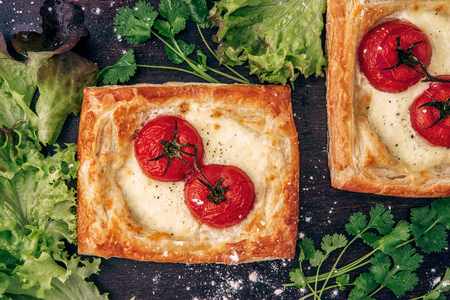 two open cheese and tomatoes pies from puff pastry with lettuce leaves and a coriander on a dark wooden background, close-upの写真素材