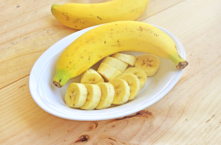 A banch of bananas and a sliced banana in a bowl over a wooden table.の写真素材