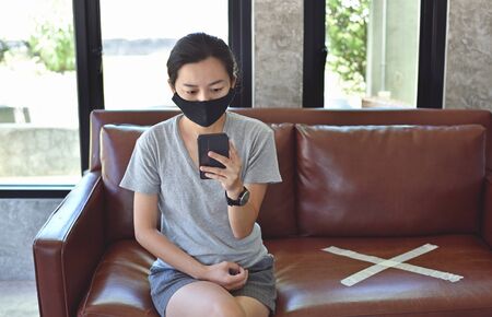 Young woman with mask using phone and sitting with distance for one seat from other people at the cafeの写真素材