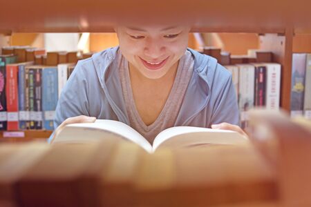 Young Asian woman university student reading book sitting by bookshelf in college library for educationの写真素材