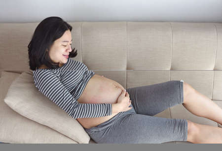 Happy smiling pregnant woman laying on couch and touching her belly at homeの写真素材