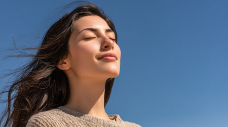 A serene young woman stands outdoors under a clear blue sky, enjoying the warmth of the sun while expressing joy and calmness, perfect for themes of relaxation and wellbeing.の素材