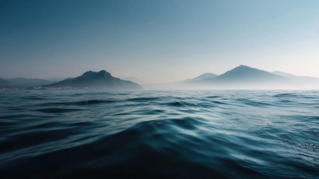 A breathtaking view of calm ocean waters with gentle ripples reflecting the light of the early blue sky. Misty mountains rise gracefully in the background, creating a serene atmosphere.の素材