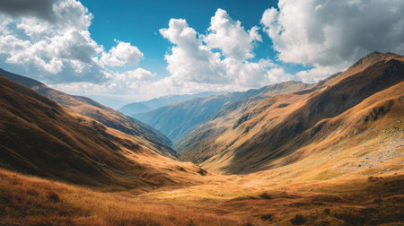 Experience the stunning beauty of a mountain valley adorned with golden grasses and towering peaks under a bright blue sky with fluffy clouds, showcasing natureの素材