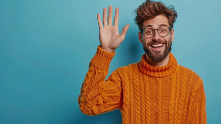 On a blue backdrop, a young, beautiful man with a beard is casually clothed with glasses and a sweater. Salutations with a grin, a wave, and a warm welcomeの素材