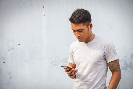 Handsome young man is staring at his cell phone against an old wall for background. Selective focus.の写真素材