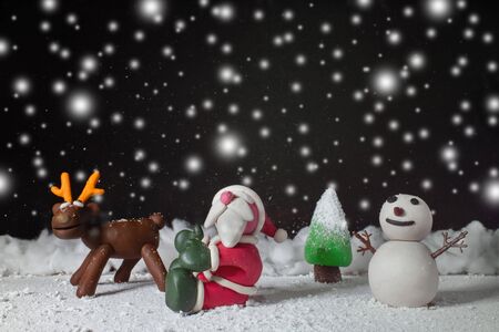 Statues like cute Santa Claus, Reindeer, Snowman and Pine trees in the snowy atmosphere for children. Molding from plasticine. Cartoon character for Christmas season on background is dark with snow at nitght.の写真素材