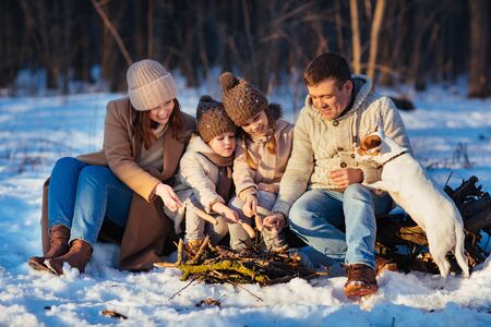 Family with two children, daughters, and dog sitting by the fire in winter snowy forest and roast on the fire sausageの写真素材