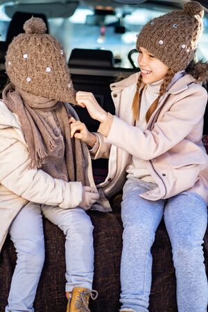 Two little sisters in winter clothes sitting in the trunk of a car in the winter and playの写真素材