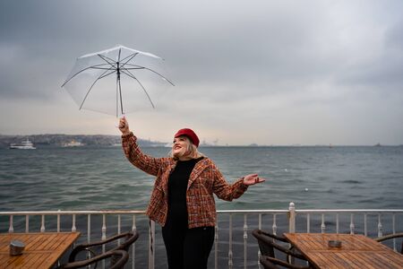 a girl with an umbrella and a red cap stands in the rain by the sea between the cafe tablesの写真素材
