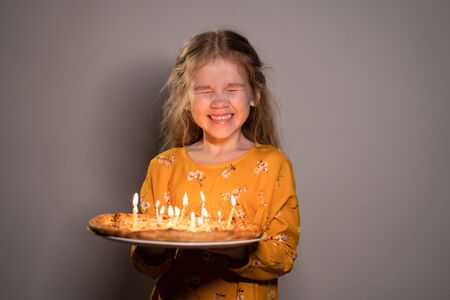 a little blonde girl holding a pizza with candles, laughing with her eyes closed on a white...