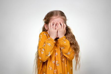 The little girl is afraid and covers her face with her hands. In front of a white wall. Girl with...