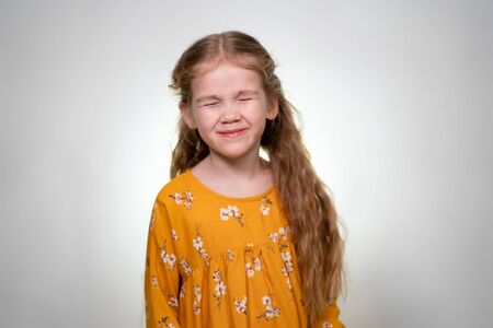 The little girl smiles with closed eyes. In front of a white wall. Girl with long hair in a yellow dressの写真素材