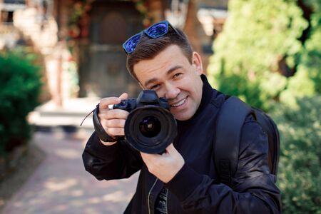 A male photographer in black clothes and sunglasses with a camera in his hands smiles