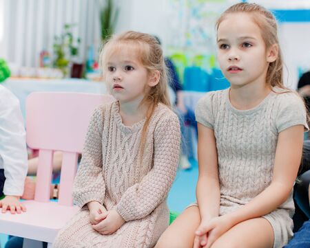 two little blonde girls in knitted dresses sit on chairs and watch the performance carefullyの写真素材