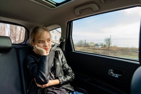 sad teenager girl in a black leather jacket rejoices looks out the window in the back seat of the...