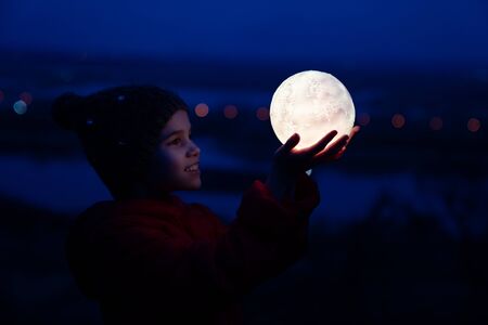 teenager girl in a cap and a red jacket holds a full glowing moonの写真素材