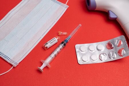 disposable medical mask, pills, syringe and non-contact thermometer on a red background. isolated...