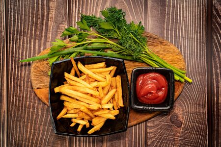 a potatoes fries in a black dish, greens  and two sauces red and white. close-up. wooden Board.