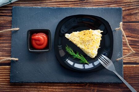a triangular piece of omelet on black plate and a tray with ketchup. the wooden table. home-cooked food.の写真素材