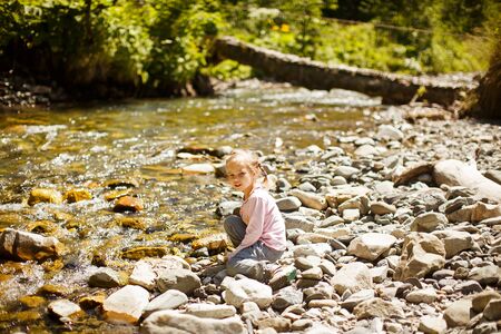 little girl in a pink sweater and jeans sitting on the rocks on the shore of a mountain river in the forest. family trip.の写真素材