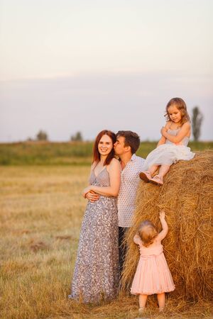 family with daughters near haystacks on a sloping field. summer sunset in the village.の写真素材