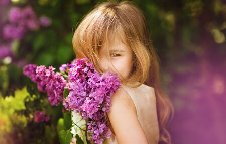 beautiful little girl in a pink dress near a bush of lilac flowers with a bouquet in hand. warm spring.の写真素材