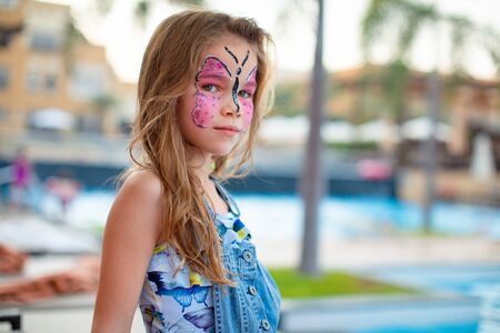 face painting pink butterfly on the face of a little girl child standing on the beach by the pool....