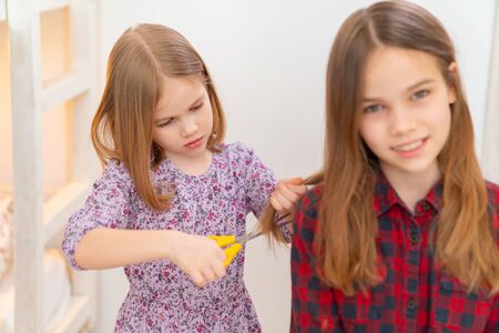 Younger sister haircut older, the children themselves make the haircut long hair with scissors, funny look, indoor portrait. haircut at home. Barber is closed.の写真素材