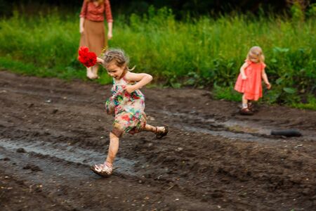 little girl in a dress jumping through puddle  in the dirty dirt road. earth after a rain. off-road. near a field with flowers poppies.の写真素材