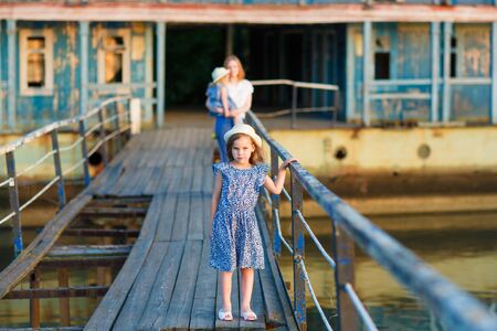 in the summer the kid girl with the mother and the sister in blue clothes standing on the old bridge broken bridge on the background of destroyed buildings. Pier on the river, naval station.の写真素材