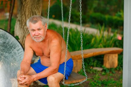 tanned elderly man sitting on the swings in his garden country house village. farming and farming in retirement.の写真素材