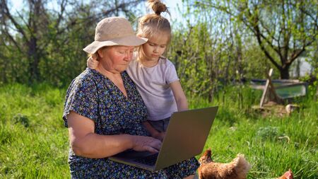 Granddaughter teaches her grandmother to working on the computer, with a laptop in village, in nature. Old woman out of town with computer. training elderly family members. the social distance.の写真素材