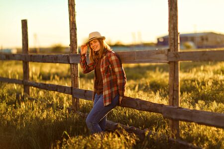 woman in hat and plaid shirt standing near a fence at sunset in the village. walks in the countryside. farming.の写真素材