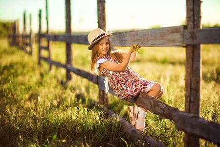 child, girl in the hat sitting on a fence in the village. walks in the countryside. farming.の写真素材