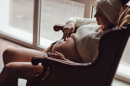 pregnant girl with a tattoo in a knitted cap sitting in the armchair in front of the window.の写真素材