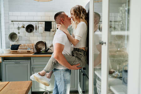 beautiful and happy pair of lovers kissing and hugging in the kitchen next to the refrigerator....
