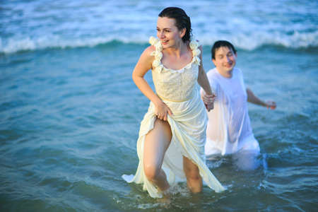 a woman leads a man by the hand in the sea water on the beach. lovers in wet clothes on their...