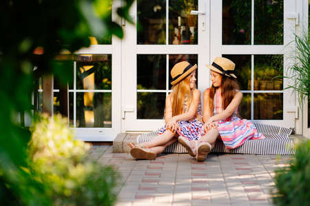 two little girls, sisters in straw hats sit on the doorstep of the house near the glass door in the back garden. children's friendship in the family.の写真素材