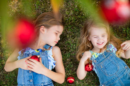 little girls with long hair in denim overalls lie on the grass under a palm tree with red Christmas toys. happy carefree childhood. new year in the tropics.の写真素材