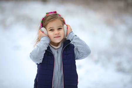 a little girl in warm headphones walks in the winter in a snowy Park and holds her head with her...