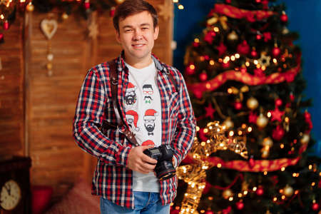 male photographer in a plaid shirt with a camera at the Christmas tree. traditional New year family...