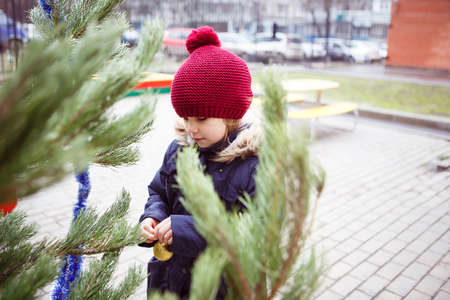 a little girl in a red hat stands near a pine tree and hangs a toy on a branch. Christmas tree in the courtyard of an apartment building.の写真素材