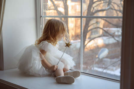 a little girl in a smart, fluffy white dress with a magic wand in her hands near the window and waits for Santa Claus or the tooth fairy. children's fairy tales and dreams. new year holiday.の写真素材