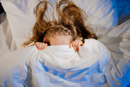 top View. Little girl lies in bed, frightened and covers face with blanket