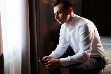 young man in a white shirt sitting on the bed and holding a bow tie