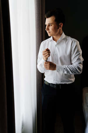 young man in a white shirt fastens cufflinks on sleeves near the windowの写真素材
