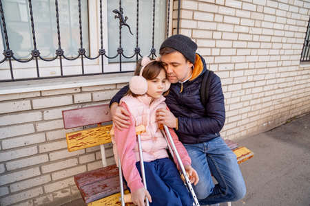 dad with daughter with crutches sits on bench in yard of apartment buildingの写真素材