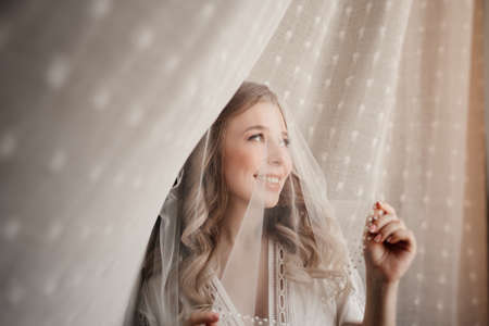 gentle morning of the bride. beautiful young woman in veil under a white curtainの写真素材
