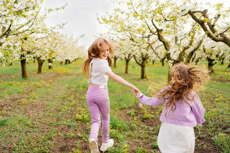 rear view beautiful and happy girls run between trees in flowering spring garden